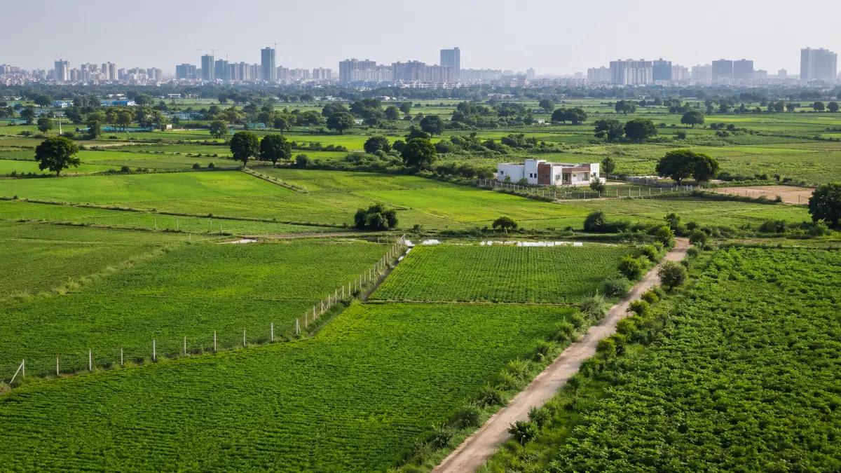 Green agricultural fields and a farmhouse near a growing Indian city